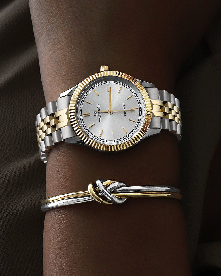 A round womens watch in silver and 22k gold from Waldor & Co. with silver sunray dial and a second hand. Seiko movement. The model is Imperial 32 Positano 32mm.